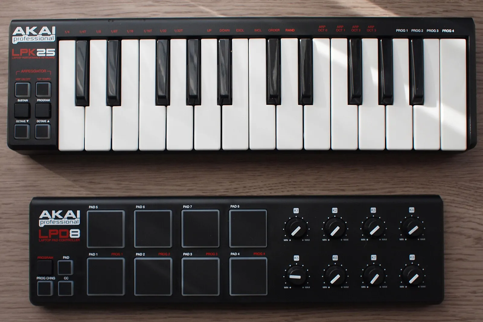 An Akai LPK25 mini keyboard above an original Akai LPD8, with its eight square pads and eight knobs, on a wooden desk.