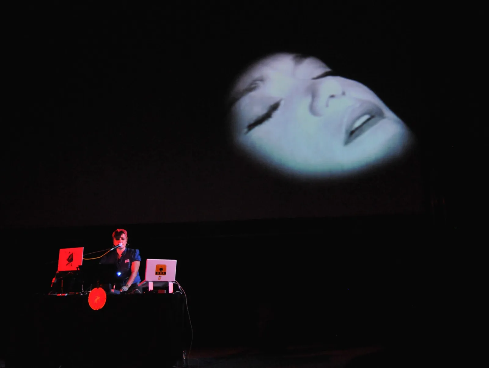 An audiovisual performance in a dark hall: a performer at a laptop lit in red on the left, and a huge projected face with closed eyes floating above.