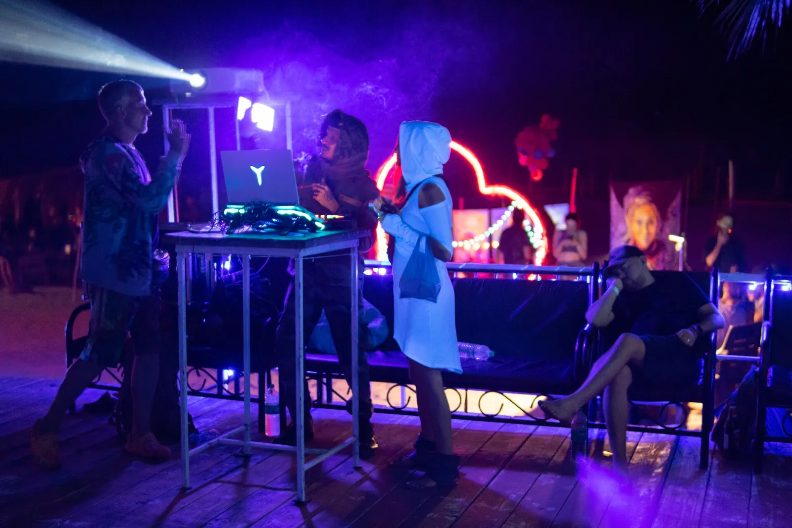 A VJ and DJ booth at an outdoor trance party at night, lit purple, with people standing around a table of equipment and a projection behind.