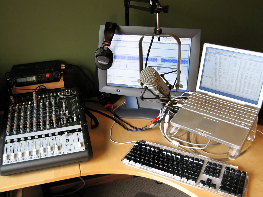 A home studio desk: a small mixer, a microphone on a boom arm, headphones, a monitor with an audio waveform and a laptop.