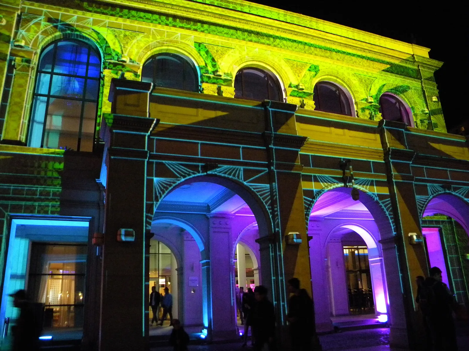A classical building facade at night covered in projected pink, purple and green light, with columns and arches picked out, and a crowd below.