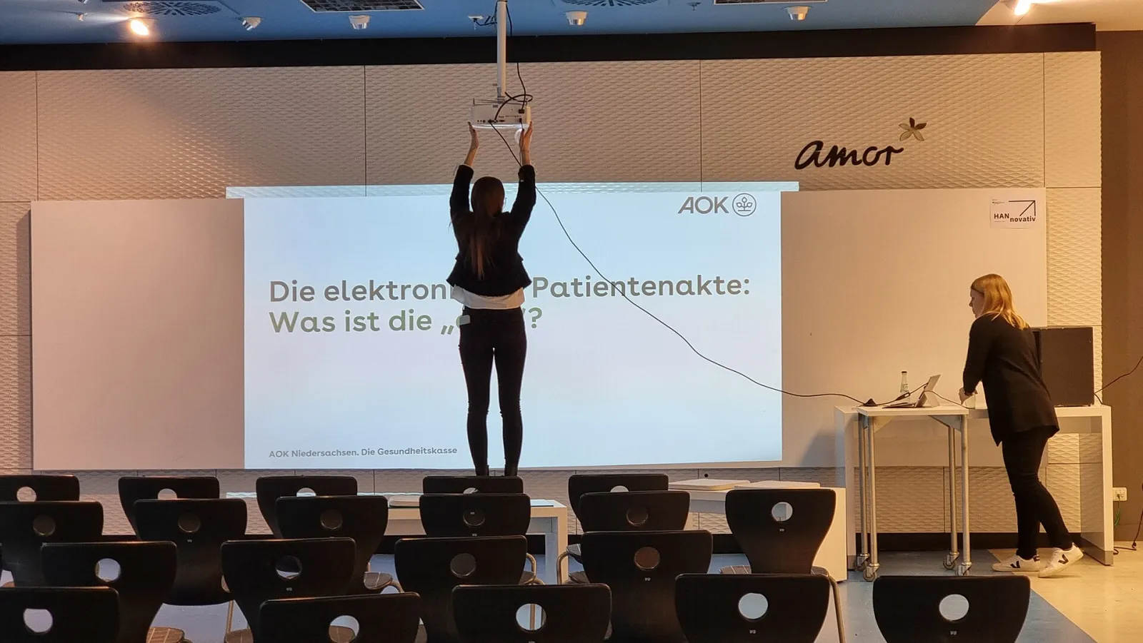 An event hall with a projection screen on stage, a person adjusting a projector hanging above the rows of seats before a talk.