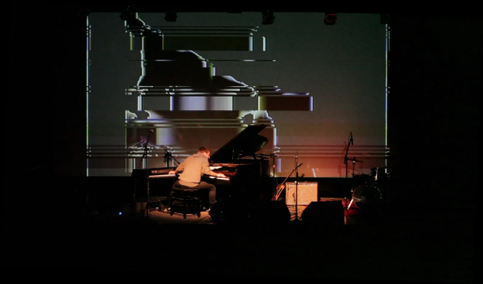 A dark stage with a grand piano and a performer, lit by a large projected image on a screen behind them.