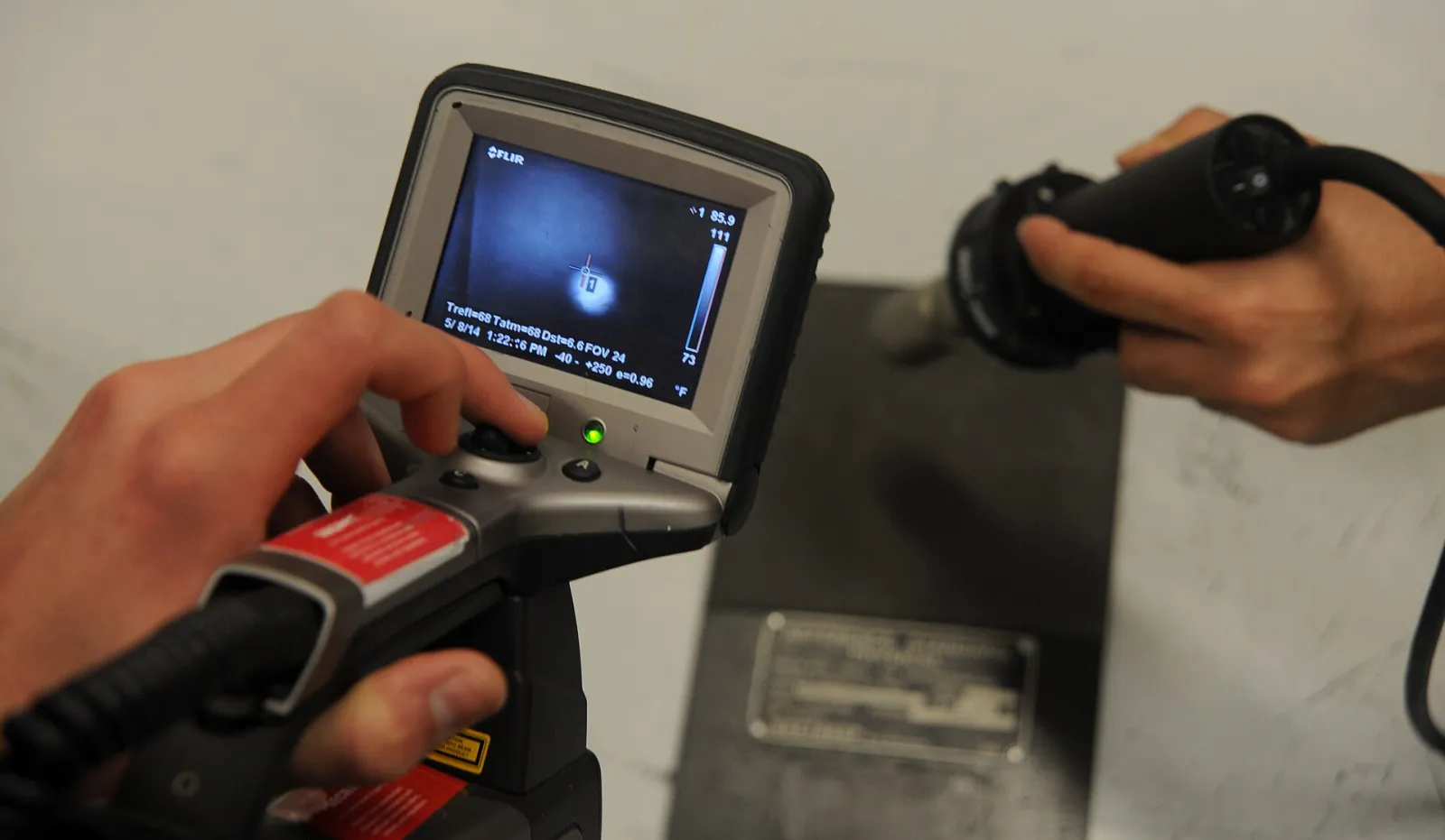 A technician holding a handheld thermal camera with a small screen, pointing it at a metal aircraft part in a workshop.